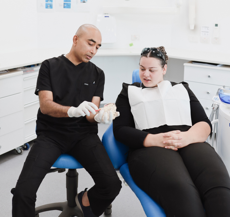 Whitehills Dental Practice Smiling woman sitting in a dental chair with a pink dental bib, showing her white teeth. A dentist wearing a white coat and face mask is visible in the background.